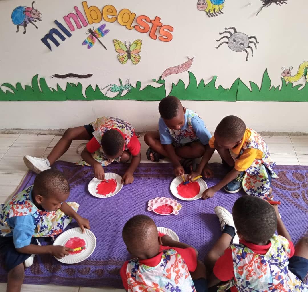 Nursery children painting paper plates on the floor beneath the minibeasts display